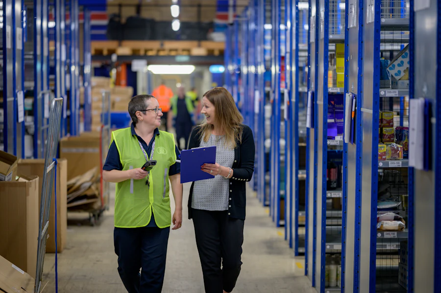 Two warehouse staff walking between blue shelving with a clipboard